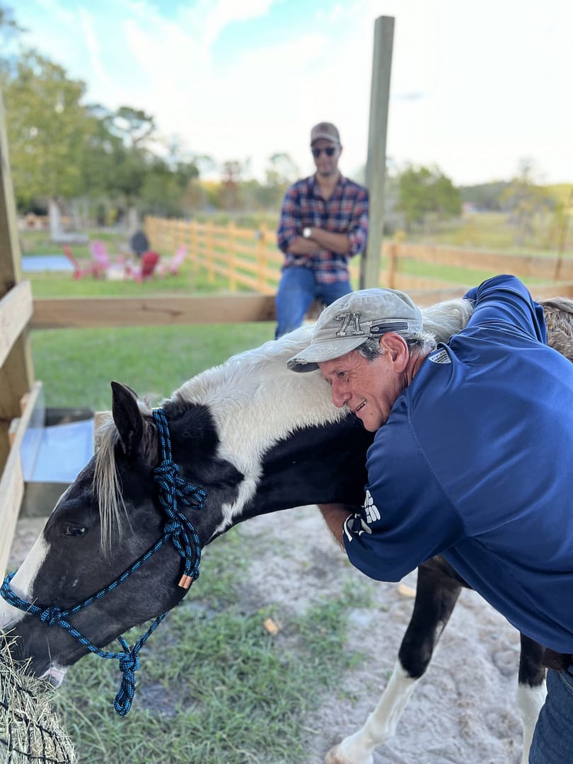 An older man in a blue shirt hugging a brown and white horse while another man in a plaid shirt and cap watches in the background at Golden Creek Ranch in Florida.