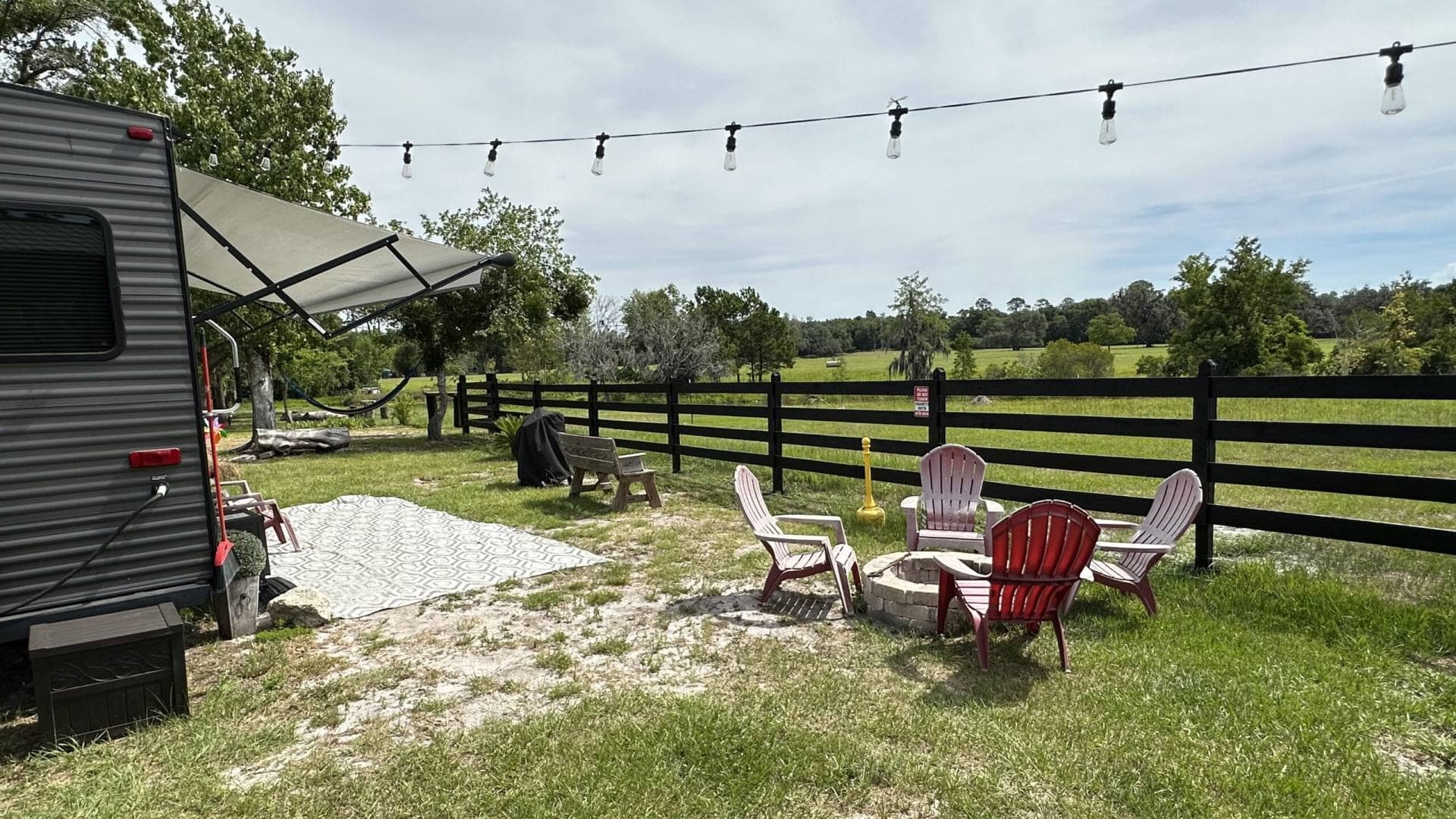 Outdoor seating area with Adirondack chairs around a fire pit, a hammock in the background, and a trailer with an awning in a scenic countryside setting in Florida